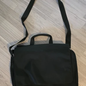 Svart datorväska med axelrem - Säljer en praktisk svart datorväska med justerbar axelrem. Väskan har flera fack och kan öppnas från både framsida och baksida. Perfekt för att bära med sig laptop och andra viktiga saker till jobbet eller skolan.