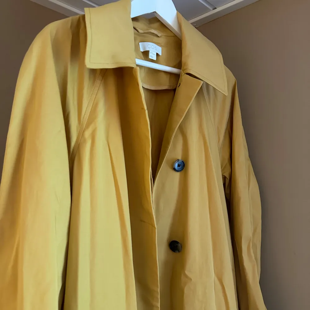 Säljer en superfin gul trenchcoat från COS i storlek M. Den har en klassisk design med stora knappar och långa ärmar. Perfekt för vår och höst när du vill ha något stilrent och bekvämt. Jackan är i bra skick och redo för nya äventyr!. Jackor.