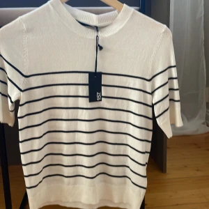 Vit och marinblå randig tröja från 157 - Säljer en superfin vit tröja med svarta ränder från märket lager  157. Den är stickad och har korta ärmar, perfekt för vår och sommar. Tröjan är i storlek S och har en klassisk rund halsringning. Den är i nyskick och har fortfarande prislappen kvar. Perfekt för en casual look eller en dag på stan!