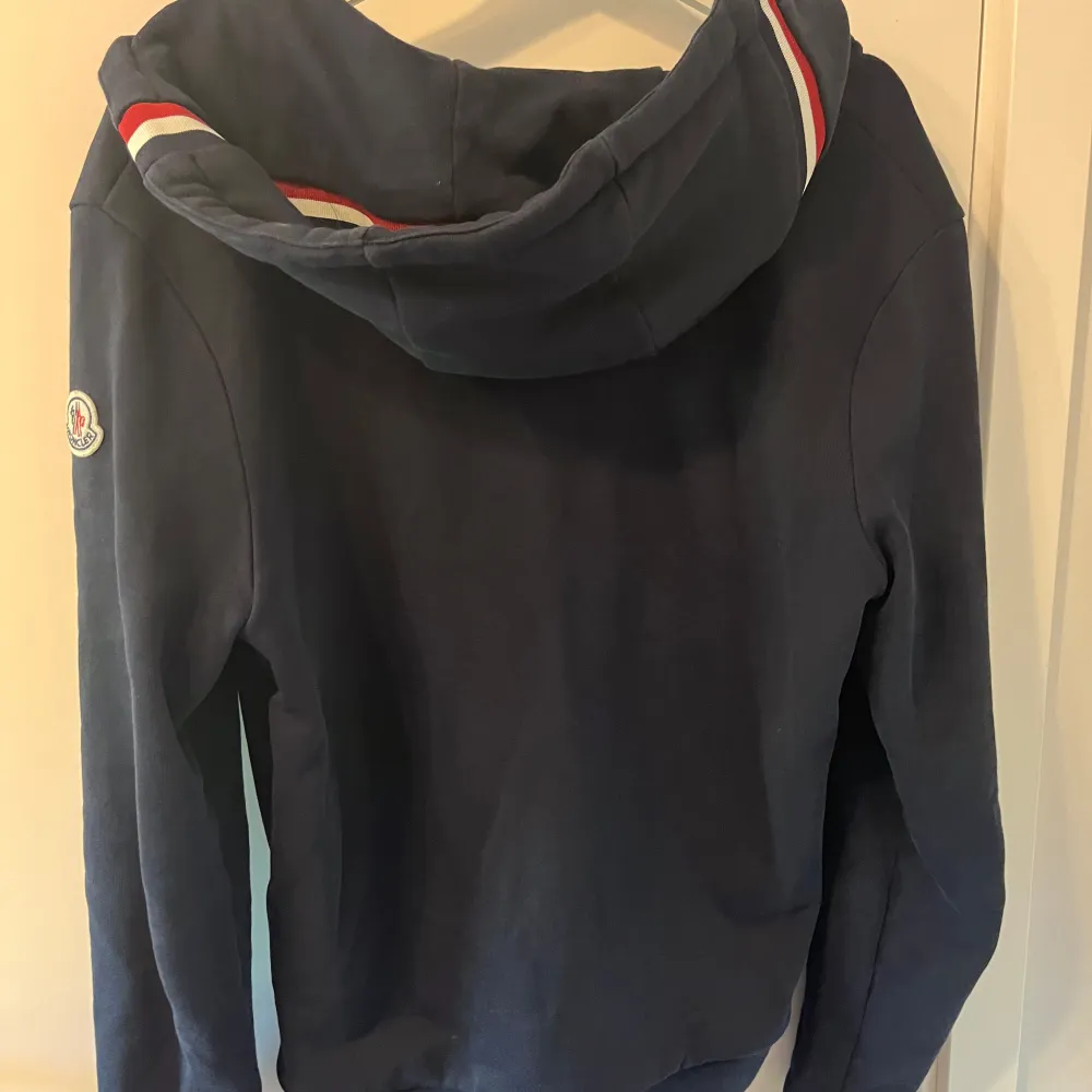 Säljer en snygg och stilren mörkblå hoodie från Moncler. Den har en dragkedja framtill och en klassisk huva med snören. På baksidan av huvan finns en röd och vit randig detalj. Monclers logga är broderad på ärmen. Skriv vid minsta fråga😁. Tröjor & Koftor.