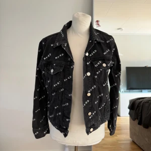 Svart jeansjacka från NA-KD - Säljer en snygg svart jeansjacka från NA-KD i storlek 34. Jackan har ett coolt mönster med NA-KD-loggan över hela tyget och silverfärgade knappar framtill. Perfekt för en casual look eller att piffa upp en outfit. Den är långärmad och har en klassisk krage.