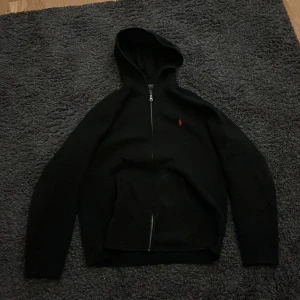 Svart huvtröja från Ralph Lauren - Säljer en stilren svart huvtröja från Ralph Lauren med dragkedja och broderad logga i rött på bröstet. Tröjan har långa ärmar och en praktisk huva. Xl barn passar s. 9/10 skick