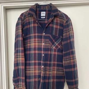 Rutig flanellskjorta från Zara - Säljer en snygg rutig skjorta från Zara i mörkblått med inslag av rött och gult. Skjortan har långa ärmar och en bröstficka. Perfekt för en avslappnad stil med sin relaxed fit. Knappar framtill och på ärmsluten.