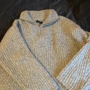 Grå stickad tröja  - Säljer en mysig grå stickad tröja med dragkedja och hög krage. Perfekt för kyliga dagar med sin ribbade design och långa ärmar. En stilren och bekväm tröja för alla tillfällen.