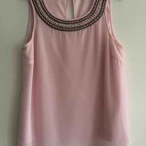 Rosa ärmlös topp från Vila - Säljer en söt rosa ärmlös topp från Vila med dekorativa pärlor och paljetter runt halsringningen. Toppen har en lätt och luftig design, perfekt för varma dagar. Den har en liten öppning i ryggen som ger en extra detalj.