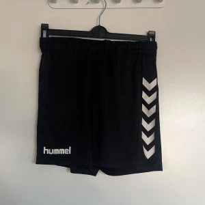 Svarta shorts från Hummel - Snygga svarta shorts från Hummel med klassiska vita pilar på sidan. Perfekta för sport och träning. Elastisk midja för bekväm passform.