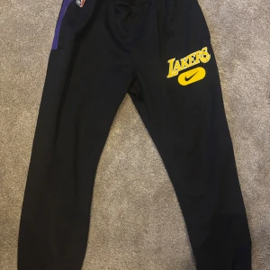 Svarta Lakers träningsbyxor från Nike - Snygga svarta träningsbyxor från Nike med Lakers-logga i gult och lila. Byxorna har en elastisk midja och en lila dragkedja på sidan. Perfekta för basketträning eller chill.