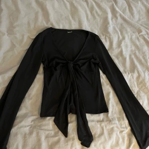 Svart långärmad topp från Gina Tricot - Snygg svart långärmad topp från Gina Tricot med knytning framtill. Perfekt för en stilren look. Toppen har en v-ringad design och är tillverkad i ett mjukt material.