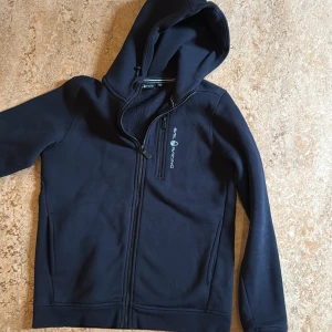 Svart huvtröja från Sail Racing - Säljer en svart huvtröja från Sail Racing med dragkedja och en bröstficka. Tröjan har en bekväm huva och långa ärmar, perfekt för kyliga dagar. Den är stilren och enkel att matcha med andra plagg. Passar både för vardag och fritid.