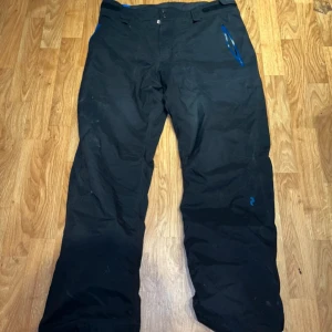 Svarta täckbyxor från Peak Performance - Svarta täckbyxor från Peak Performance med blå detaljer och knappar i midjan. Perfekta för vinterns kalla dagar. Byxorna har en loose passform och är tillverkade i ett slitstarkt material.