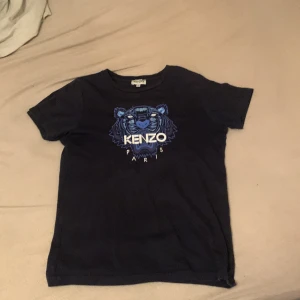 Svart t-shirt med tigertryck från Kenzo - Säljer en svart t-shirt från Kenzo med ett coolt tigertryck i blått och vitt på framsidan. T-shirten är kortärmad och har en rund halsringning. Perfekt för dig som vill ha en stilren men ändå iögonfallande look. 🐯