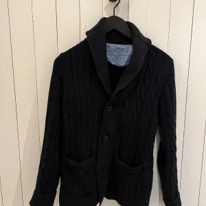 Stickad cardigan  - Snygg svart stickad cardigan från Gant med kabelstickat mönster. Koftan har långa ärmar och knappar framtill samt två fickor. Perfekt för kyliga dagar.