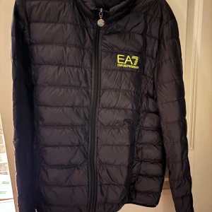 Svart pufferjacka från EA7 - Säljer en svart pufferjacka från EA7 Emporio Armani med dragkedja framtill. Jackan har en quiltad design och långärmade ärmar. Perfekt för kyligare dagar.