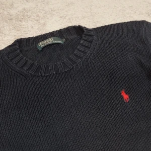 Svart stickad tröja från Ralph Lauren - Säljer en klassisk svart stickad tröja från Ralph Lauren med det ikoniska röda logotypbroderiet på bröstet. Tröjan har en rund halsringning och ribbade muddar vid ärmslut och nederkant. Perfekt för en stilren look.