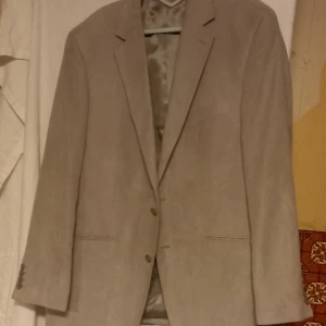 Beige kavaj från Dressmann - Säljer en stilren beige kavaj från Dressmann. Kavajen har en klassisk design med två knappar framtill och är tillverkad i ett mjukt material. Perfekt för både formella och semi-formella tillfällen. Fodret är i en matchande beige ton.