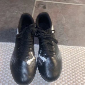 Svarta fotbollsskor från Puma  - Snygga svarta fotbollsskor från Puma med vit logga och snörning. Perfekta för spel på konstgräs med sin slitstarka sula och stilrena design.