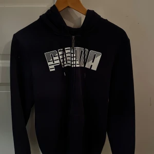 Svart hoodie från Puma - Säljer en svart hoodie från Puma med dragkedja och stor vit logga på framsidan. Perfekt för en avslappnad stil. Den har en bekväm huva med snörning och långa ärmar.