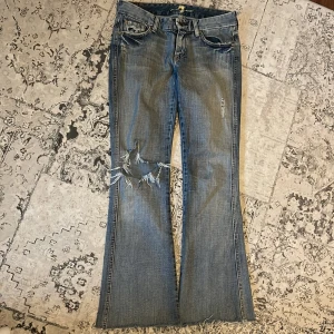 Blå bootcut jeans från 7 for All Mankind - Snygga blå bootcut jeans med slitningar på ena benet och coola detaljer på bakfickorna. Perfekta för en avslappnad stil med en touch av edge. Märket 7 for All Mankind garanterar kvalitet och stil.