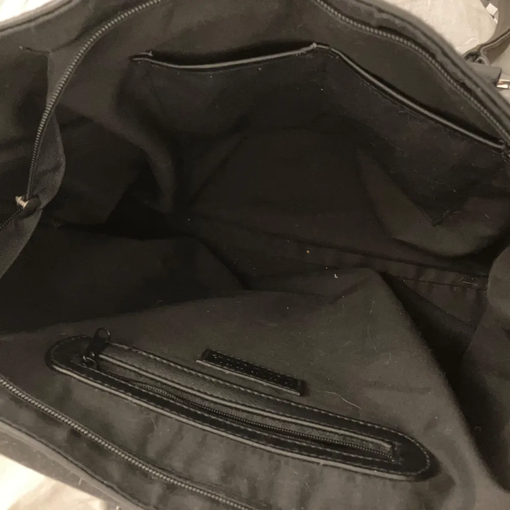 Snygg svart axelväska i syntetmaterial med praktisk dragkedja och stiliga metallspännen. Väskan har ett rymligt inre med flera fack för enkel organisering. Perfekt för att bära med sig allt man behöver under dagen.. Laukut & Käsilaukut.
