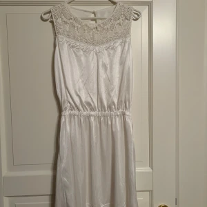 Vit klänning med spetsdetaljer - En vit klänning med spets upptill och ärmlös design. Materialet är sidenliknande och glansigt. Klänningen har en elastisk midja som framhäver figuren fint. I ryggen en knapp. Lår- knälång. Sällan använd.
