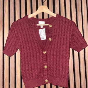 Röd stickad kofta från H&M - Säljer en snygg röd stickad kofta från H&M med korta ärmar och dekorativa guldknappar. Koftan har ett klassiskt flätmönster och ribbade kanter.