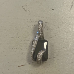 Silverfärgat hänge med svart sten - Elegant hänge i silverfärg med en stor svart sten. Stenen är omsluten av en böljande design med små glittrande detaljer. Perfekt för att ge en stilren touch till din outfit.