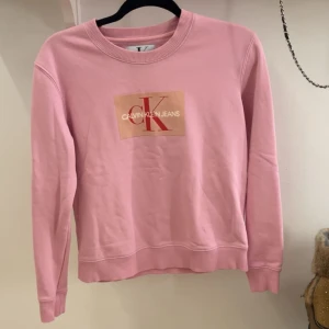 Rosa sweatshirt från Calvin Klein - Snygg rosa sweatshirt från Calvin Klein med deras ikoniska logga på bröstet. Tröjan har en rund halsringning och långa ärmar, perfekt för en avslappnad stil. Passar bra till jeans eller leggings för en casual look.