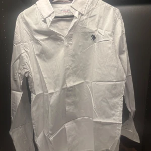 Vit skjorta från U.S. Polo Assn. - Säljer en klassisk vit skjorta från U.S. Polo Assn. med broderad logga på bröstet. Skjortan har långa ärmar och knappar framtill. Perfekt för en stilren look.