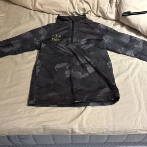 Grå camouflagemönstrad tröja med dragkedja - Säljer en grå camouflagemönstrad tröja från ATH DEPT. Tröjan har en halv dragkedja framtill och långa ärmar. Perfekt för en sportig look eller avslappnad stil.