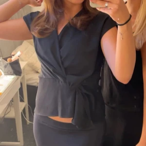 Svart omlottopp - ! Inte mina bilder!!!Snygg svart omlottopp med knytning i midjan. Toppen har en elegant v-ringad design och är ärmlös, vilket gör den perfekt för både vardag och fest. Passar bra till jeans eller kjol för en stilren look.