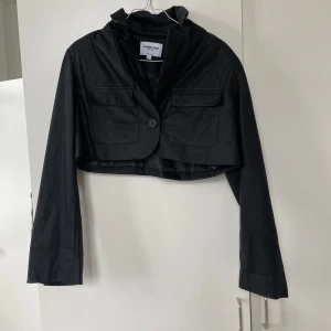 Svart croppad jacka från NA-KD - Säljer en snygg svart croppad jacka från NA-KD i samarbete med Romee Strijd. Jackan har en knapp framtill och två fickor på bröstet. Perfekt för en cool och stilren look. Passar bra till både vardag och fest!