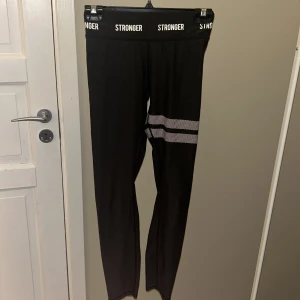 Svarta träningsbyxor från Stronger - Säljer ett par svarta leggings från Stronger i mycket bra skick. De har en elastisk midja med logotypen 'STRONGER' och en vit randdetalj runt låret. Perfekta för träning eller en avslappnad dag. Materialet är stretchigt och bekvämt. Är mörkgråa, nästan svarta