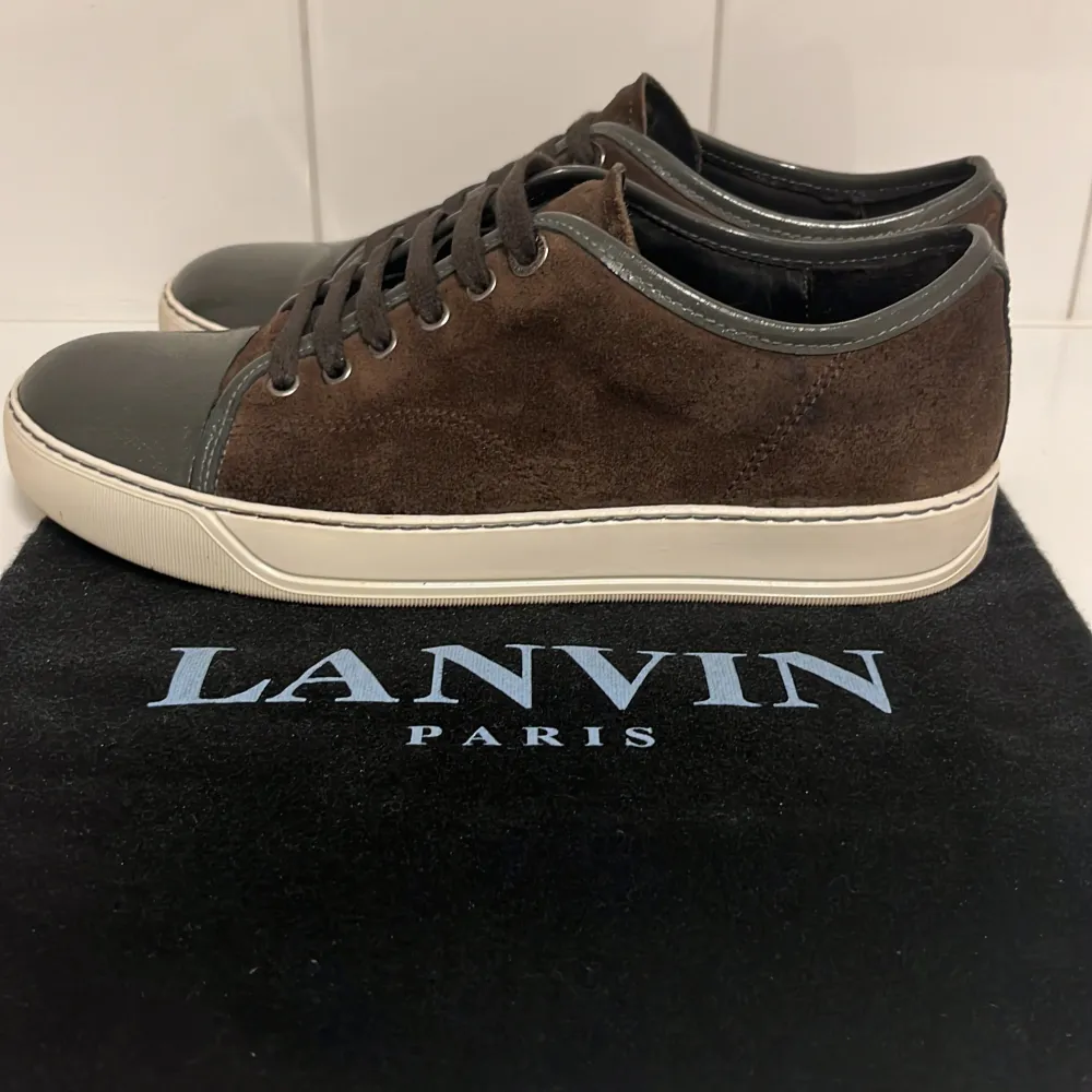 Säljer ett par snygga bruna sneakers från Lanvin i mocka med glansiga detaljer på tån och runt snörningen. De har en vit sula som ger en fräsch kontrast. Perfekta för både vardag och fest! Skorna är i mycket bra skick.. Skor.