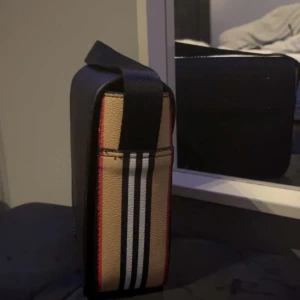 Randig axelväska - Snygg axelväska med klassiskt randigt mönster i beige, svart och vitt. Väskan har en stilren design med en svart flik som täcker öppningen. Perfekt för både vardag och fest! Burberry