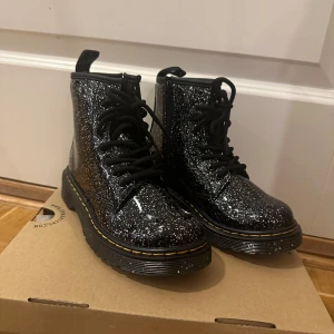 Svarta glittriga boots från Dr. Martens - Säljer ett par svarta glittriga boots från Dr. Martens i mycket bra skick. De har en robust sula och klassisk snörning. Perfekta för att ge din outfit lite extra bling! Passar både till vardags och fest.