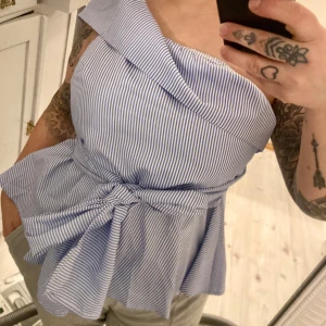 Randig topp strl 40/42 - One shoulder randig blus.  Köpt från Shein Curve. Storlek 0XL. Skulle säga att den motsvarar en 40/42.  Jag har vanligtvis storlek 40, så beroende på hur tajt man vill att toppen ska sitta så passar den både en 40 och 42. Dragkedja i sidan och knytskärp. Skärpet är inte avtagbart men kan knytas antingen fram eller bak.  Helt ny, oanvänd. 