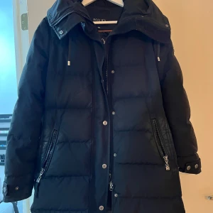 Svart dunjacka från Hollies - Säljer en snygg och varm svart dunjacka från Hollies i storlek 34. Jackan har en stilren design med dragkedjor och knappar framtill. Perfekt för kalla vinterdagar med sin långa ärm och pufferstil. Den har också en praktisk huva för extra skydd mot väder och vind. Lite glansig vid fikan men inget som märks så mycket finns ingen päls därav priset! Priset går o diskutera vid snabb affär 