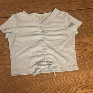 Ljusblå ribbad topp från Lindex - Säljer en söt ljusblå ribbad topp från Lindex i storlek 146/152. Toppen har korta ärmar och en v-ringad hals. Den har en snygg knytdetalj framtill som ger en fin passform. Perfekt för vår och sommar!