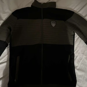 Svart och grön tröja med dragkedja - Säljer en snygg svart och grön tröja från EA7. Den har en dragkedja framtill och ett stilrent märke på bröstet. Perfekt för höst och vår med sin bekväma passform och långa ärmar. Jackan är i mycket bra skick och redo för nya äventyr!