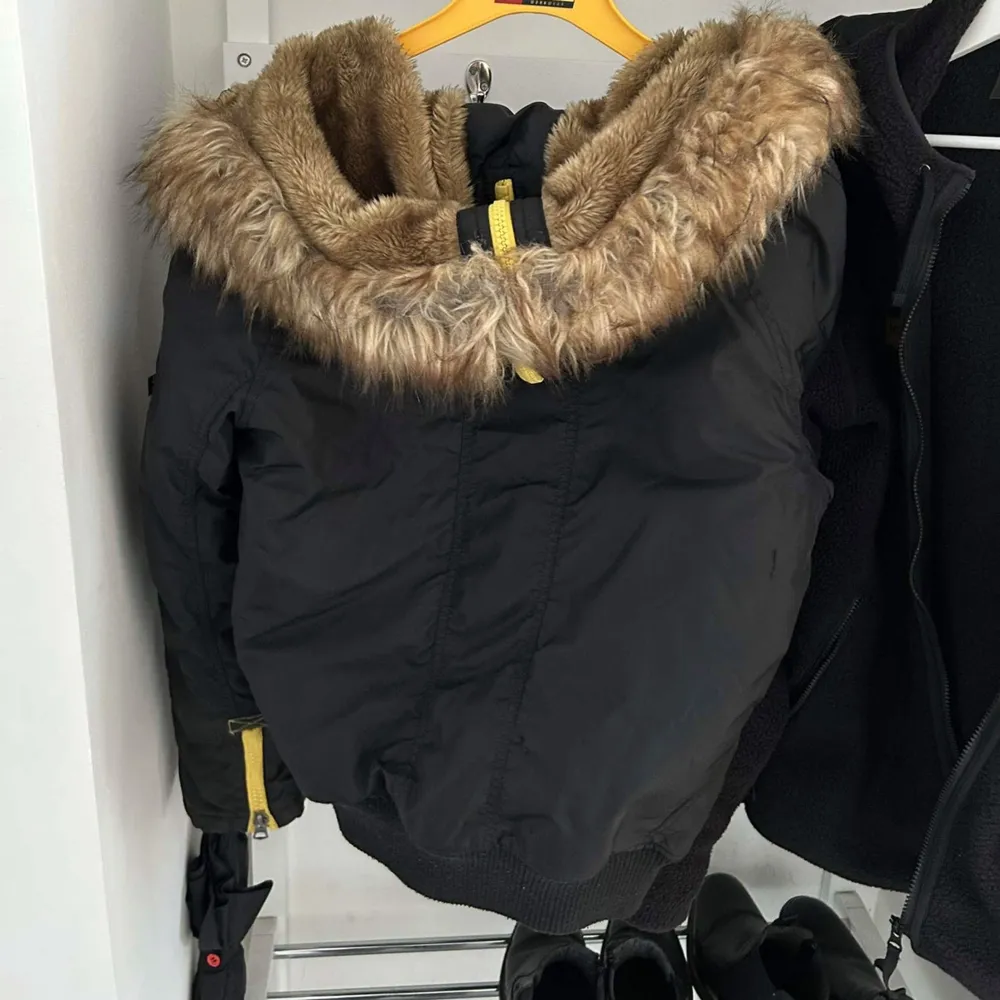 Säljer en snygg svart jacka från Alpha Industries i storlek S. Den har en mysig pälsfodrad huva och gula detaljer som ger en cool kontrast. Jackan har dragkedja och knappar framtill, samt flera fickor för praktisk förvaring. Perfekt för höst och vinter!. Jackor.