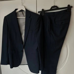Mörkblå kostym från Zara Man - Säljer en stilren mörkblå kostym från Zara Man Basic Collection. Kavajen har en klassisk passform med två knappar och byxorna är raka och eleganta. Perfekt för både formella tillfällen och fest. I mycket bra skick. Passar utmärkt för alla årstider.