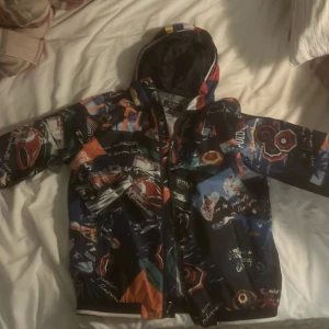 Windbreaker Polo Ralphlauren  - Säljer en cool flerfärgad jacka från Polo Ralph Lauren. Den har ett unikt mönster med olika färger och motiv, perfekt för att sticka ut. Jackan har en dragkedja och huva, vilket gör den både praktisk och snygg. Perfekt för höst och vår!