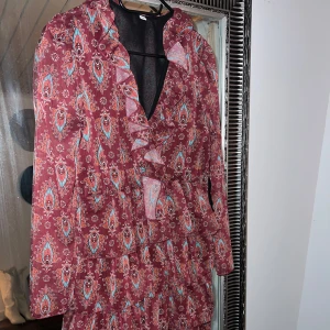 Mönstrad klänning med volanger - Säljer en superfin mönstrad klänning i vinrött med volanger. Den har långa ärmar och ett vackert paisleymönster. Perfekt för höst och vår, och funkar både till fest och vardag. Klänningen har en v-ringad design och är i mycket bra skick. Passar perfekt för dig som gillar en bohemisk stil!