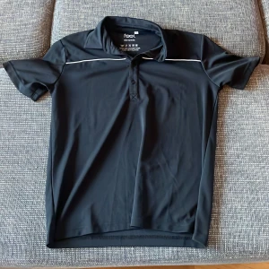 Svart kortärmad pikétröja från Feel - Säljer en stilren svart pikétröja från Feel i storlek S. Tröjan är tillverkad i 100% polyester och har en klassisk krage med knappar. Perfekt för både vardag och träning. Den har en vit linje som detalj på framsidan och baksidan. Skön och lätt att bära! Pris går alltid att förhandlas!