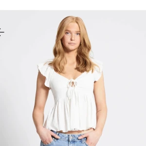 Vit blus med volangdetaljer - Söt vit blus med volangdetaljer och knyt framtill. Perfekt för sommardagar eller en avslappnad vårlook. Den är ärmlös och har en luftig passform som gör den bekväm att bära hela dagen.
