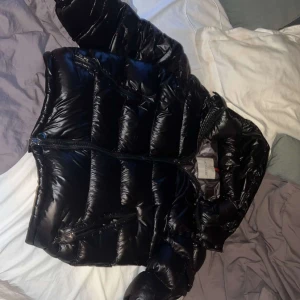 Svart pufferjacka från Moncler - Säljer en snygg svart pufferjacka från Moncler i mycket bra skick. Jackan har en glansig finish och dragkedja framtill. Perfekt för kalla vinterdagar och håller dig varm och stilren. Inuti finns en cool serietidningsdetalj som ger jackan en unik touch.