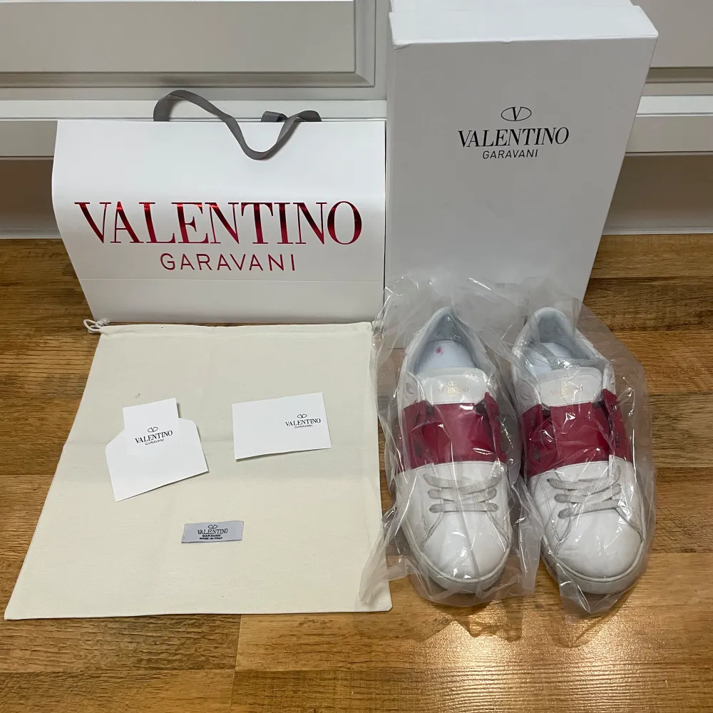 Säljer ett par snygga Valentino Garavani sneakers i vitt med en röd detalj över snörningen. Skorna är i mycket bra skick och kommer med originalförpackning och dustbag. Perfekta för både vardag och fest! Storlek är inte specificerad, men de ser ut att vara i standard herrstorlek.. Skor.