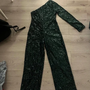 Grön paljettbyxdress  - Säljer en fantastisk grön paljettbyxdress med oneshoulder-design. Perfekt för festliga tillfällen med sin glittriga yta som verkligen fångar ljuset. Det står att den är i storlek L men jag har i vanliga fall storlek S och den är lite stor i midjan och bysten men lite kort i benen så skulle säga att den är M. Byxdressen har en elegant lång passform och är i nytt skick då jag aldrig använt den, endast testad. Säljer då den är för liten för mig. Perfekt för att göra ett statement på nästa fest!