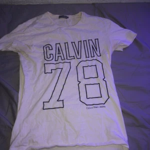 Vit t-shirt från Calvin Klein Jeans - Säljer en vit t-shirt från Calvin Klein Jeans i storlek S. Den har ett stort tryck med texten 'CALVIN 78' på framsidan. Perfekt för en avslappnad stil och passar bra till jeans eller shorts. T-shirten är i bra skick och redo för nya äventyr!
