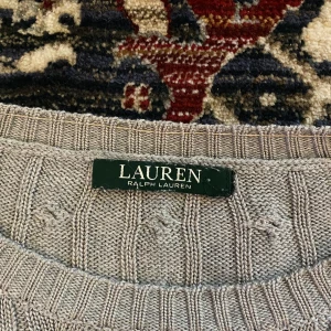 Grå kabelstickad tröja från Ralph Lauren - Säljer en stilren grå kabelstickad tröja från Ralph Lauren. Perfekt för kyliga dagar med sin mjuka och varma känsla. Tröjan har långa ärmar och en klassisk rund halsringning. Passar både till vardags och lite finare tillfällen. 🧶✨ Jag har satt dit trådar för att göra handlederna lite mindre och runt halsen, men det går enkelt att klippa bort! Annars i perfekt skick! 